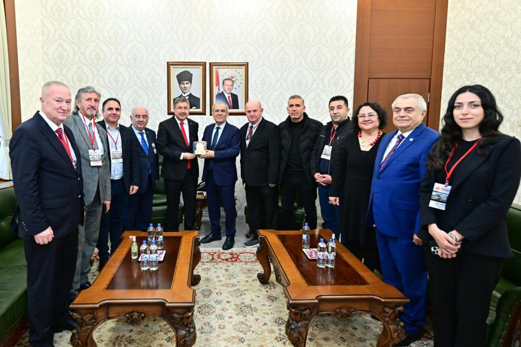 Bir grup insan, resmi bir toplantıda bir araya gelmiş. Ortada hediye alıp veren iki kişi var. Arkalarında Türkiye Cumhuriyeti'nin kurucu lideri Atatürk ve mevcut Cumhurbaşkanı'nın portreleri asılı.