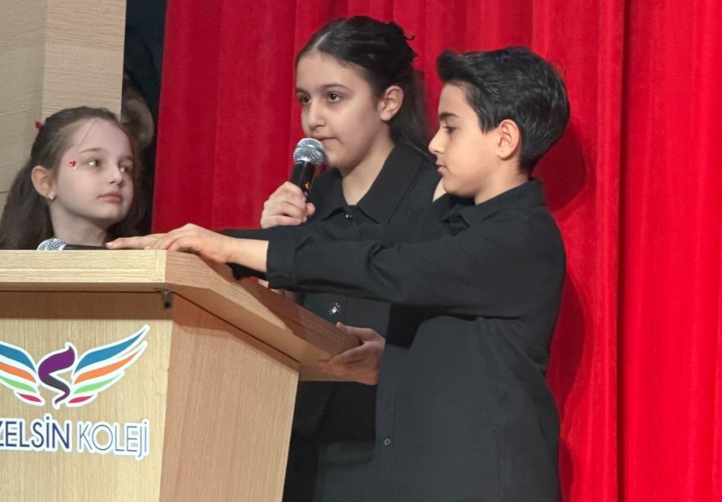 Bir podiumda konuşma yapan çocuklar, arka planda kırmızı perde ile sahnede. Bir çocuk mikrofon tutarken, diğeri onun yanında duruyor ve dikkatlice dinliyor.