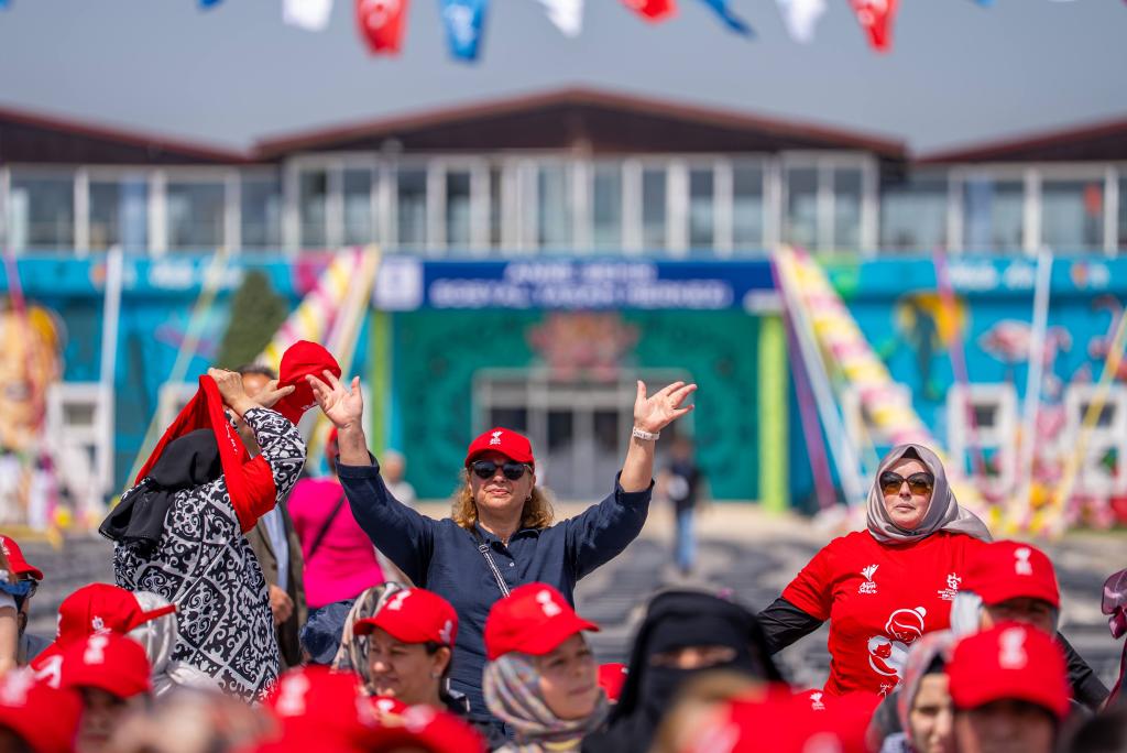Bir kalabalık etkinlikte, kırmızı şapka takan kadınlar coşkulu bir şekilde ellerini kaldırmış. Arka planda renkli bayraklar ve sanat eserleri olan bir bina yer alıyor.