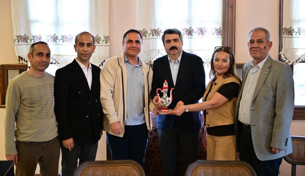 Altı kişi bir arada, biri elinde süslü bir çerçeve tutuyor. Arka planda perdeler ve pencereler görülüyor.